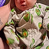 Marie is registered to the contest to win money with this photo: baby, infant, child, portrait, close_up, wide_eyed, looking_at_camera, onesie, avocado_pattern, clothing, zipper, adult_arm, tattoo, bottle, pacifier, indoor, couch, home, cozy, photography