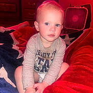 Liam is registered to the contest to win money with this photo: toddler, child, indoor, blanket, bed, long_sleeve_shirt, furniture, fan, dresser, curly_hair, sitting, blue_eyes, red_light, cozy, room, hands, portrait, person, expression, casual