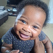 Edwin is registered to the contest to win money with this photo: baby, infant, smile, grin, close_up, portrait, polka_dot_onesie, living_room, sofa, television, cheeks, eyes, hair, hands, adult_hand, skin, cute, happy, indoor, natural_light