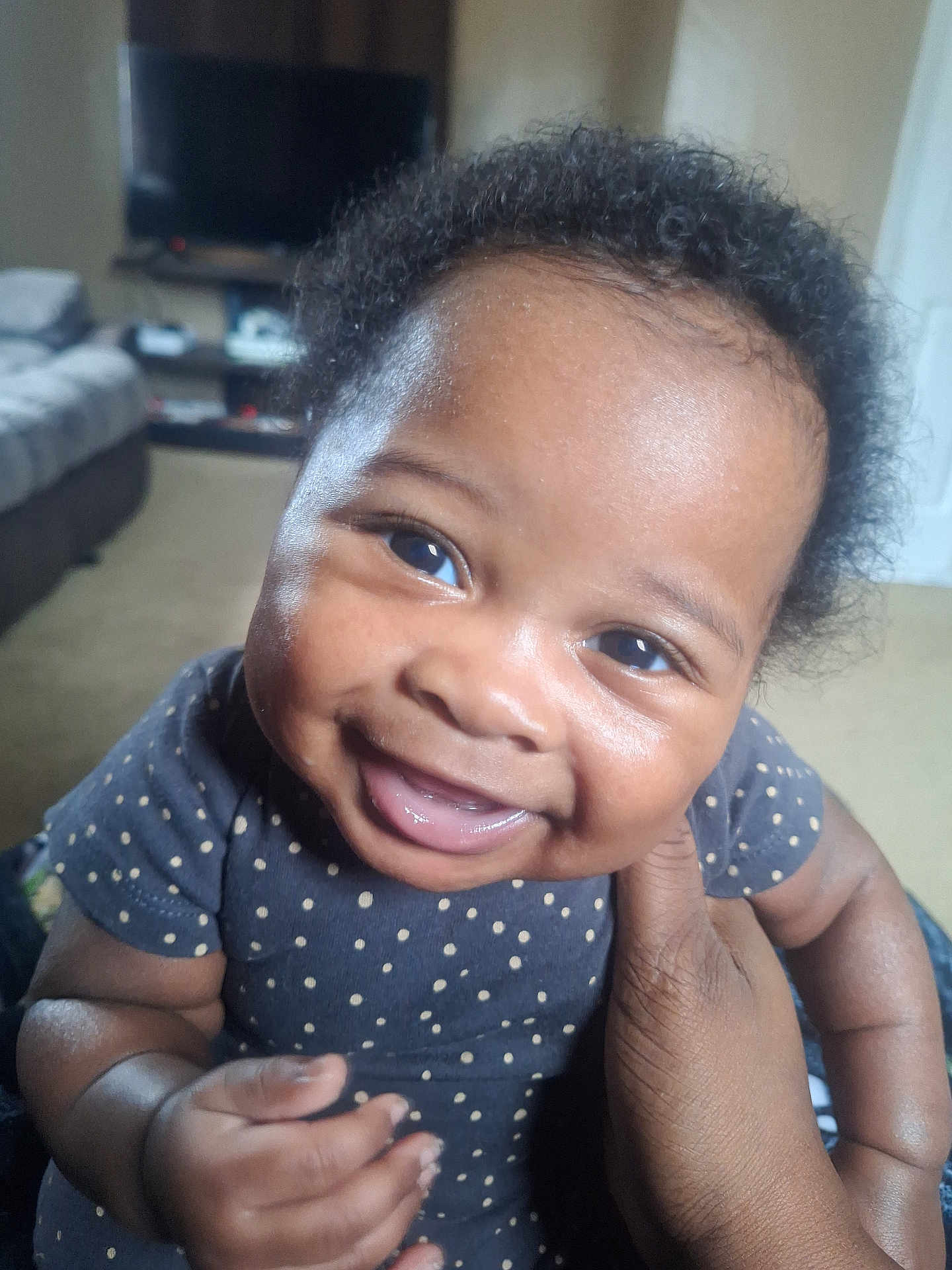 Edwin is registered to the contest to win money with this photo: baby, infant, smile, grin, close_up, portrait, polka_dot_onesie, living_room, sofa, television, cheeks, eyes, hair, hands, adult_hand, skin, cute, happy, indoor, natural_light