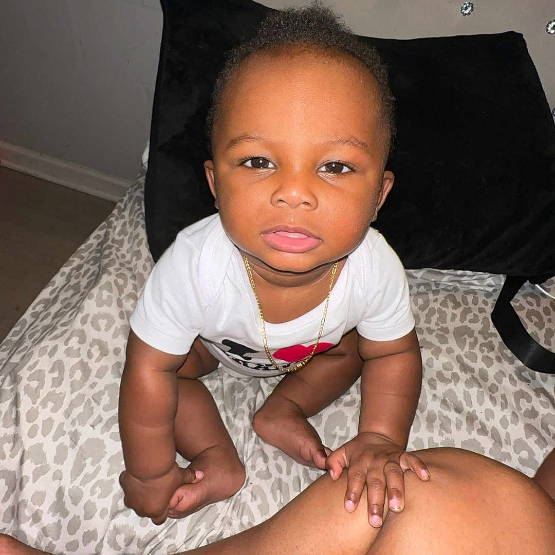 Vino is registered to the contest to win money with this photo: baby, bedspread, black_pillow, child, curious, cute, floor, gold_chain, hand, headboard, indoor, infant, leg, leopard_print, person, sitting, skin, studs, touching, white_shirt
