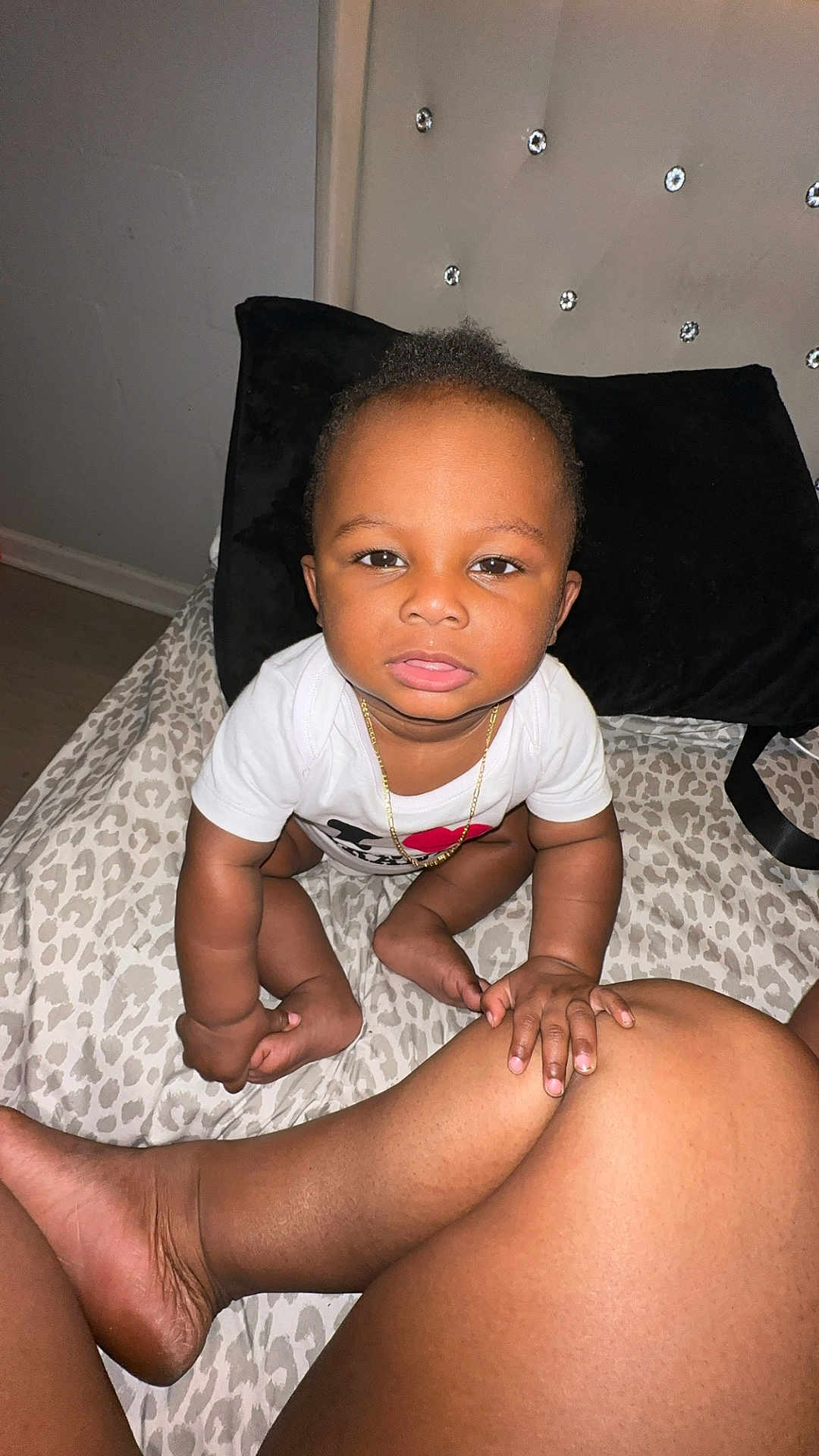 Vino is registered to the contest to win money with this photo: baby, child, infant, white_shirt, gold_chain, leopard_print, bedspread, black_pillow, headboard, studs, hand, leg, skin, indoor, floor, person, cute, curious, sitting, touching