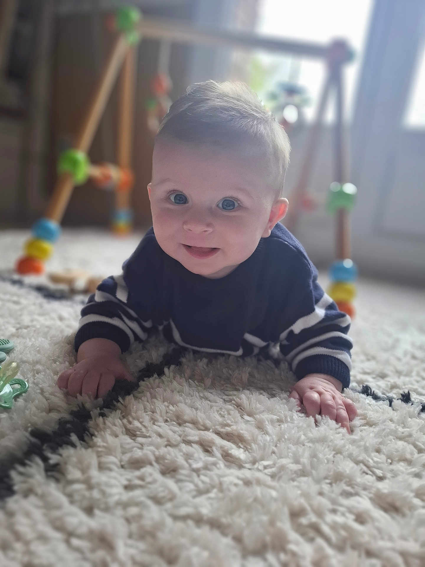 Julian a rejoint le concours — aidez-le/la à gagner de superbes lots ! baby, infant, crawling, rug, carpet, toy, wooden_toy, colorful, blue_eyes, striped_sweater, indoor, floor, child, cute, smiling, hand, face, person, young_child, playtime