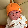 Tommy is registered to the contest to win money with this photo: baby, infant, hat, orange_hat, pumpkin, onesie, cartoon_print, face, portrait, cushion, beige_background, clothing, cute, indoors, child, newborn, expression, headwear, sitting, closeup