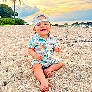 Landon is registered to the contest to win money with this photo: baby, beach, sand, sunset, ocean, palm_tree, child, smiling, cap, tropical_print, nature, outdoor, vacation, happy, sitting, footwear_free, sky, coast, toddler, casual_clothing