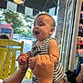 Matthias J is registered to the contest to win money with this photo: baby, child, striped_clothing, hands, yellow_chair, table, window, indoor, smiling, happy, person, face, daylight, phone, tattoo, brick_wall, shelf, candy, store, playful