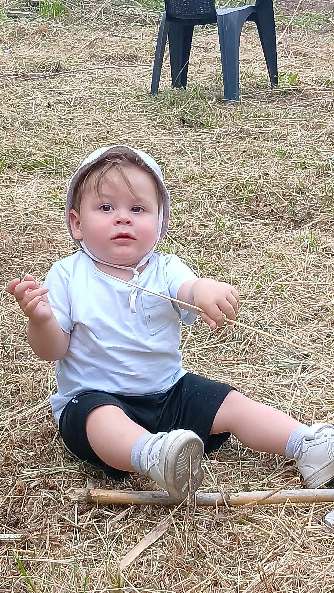 Kenzo a rejoint le concours — aidez-le/la à gagner de superbes lots ! toddler, child, sitting, grass, outdoor, hat, white_tshirt, black_shorts, stick, shoes, socks, plastic_chair, nature, curious, innocent, daylight, casual_clothing, young_child, ground, playful