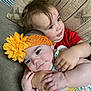 baby, child, hug, couch, pillow, flower_headband, red_shirt, dress, indoor, closeup, toddler, affection, cute, siblings, family, love, comfort, skin, face, hands
