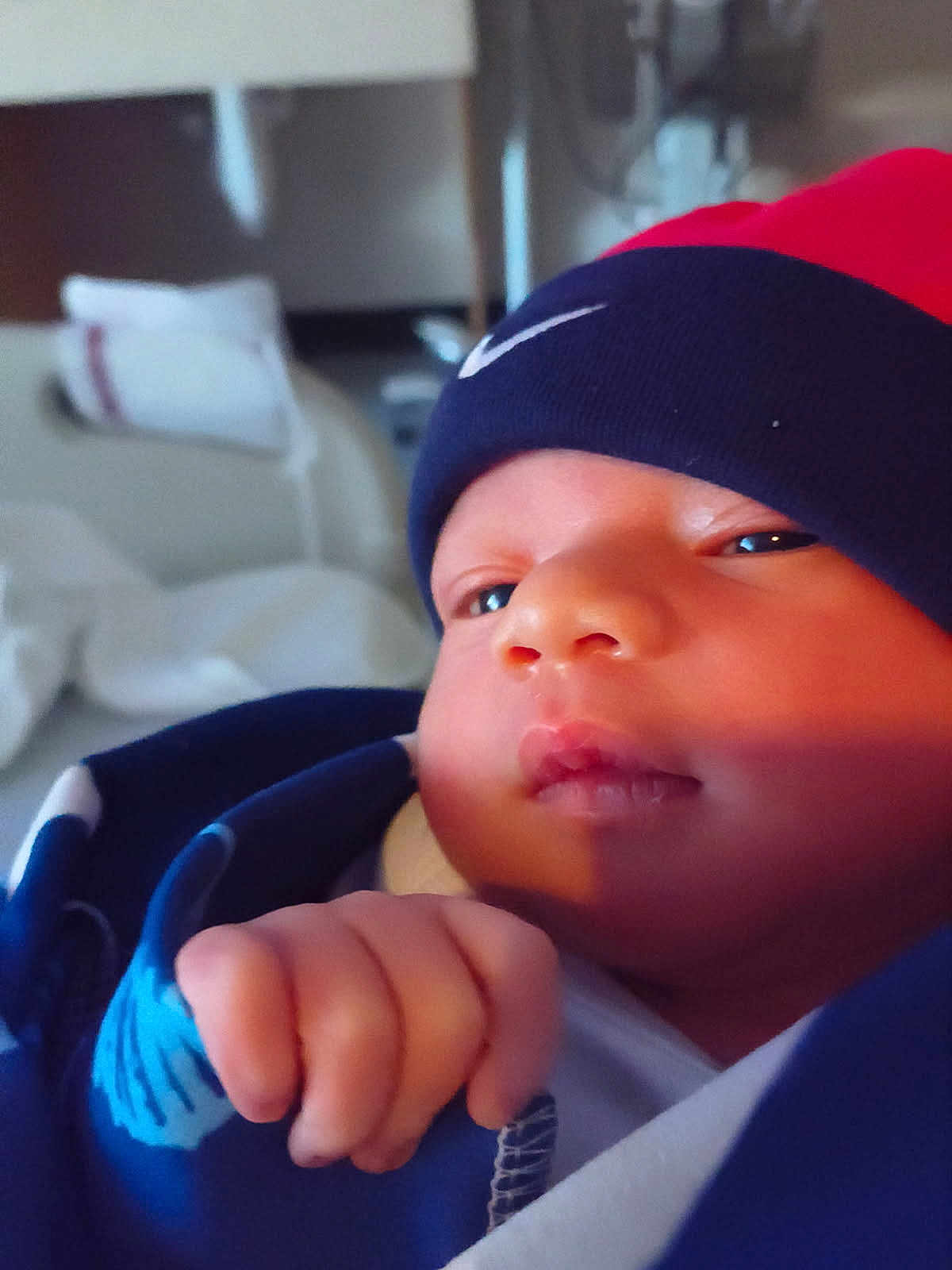Tevon Xavier Brown is registered to the contest to win money with this photo: baby, infant, close_up, hat, blue_blanket, red_hat, face, hand, fist, indoors, soft_lighting, portrait, newborn, clothing, person, cute, skin, expression, wrapped, cozy
