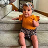 baby, child, infant, carpet, stairs, headband, orange_clothing, leopard_print, sitting, indoors, smiling, person, portrait, cute, home_interior, shelf, bag, floor, wall, foot