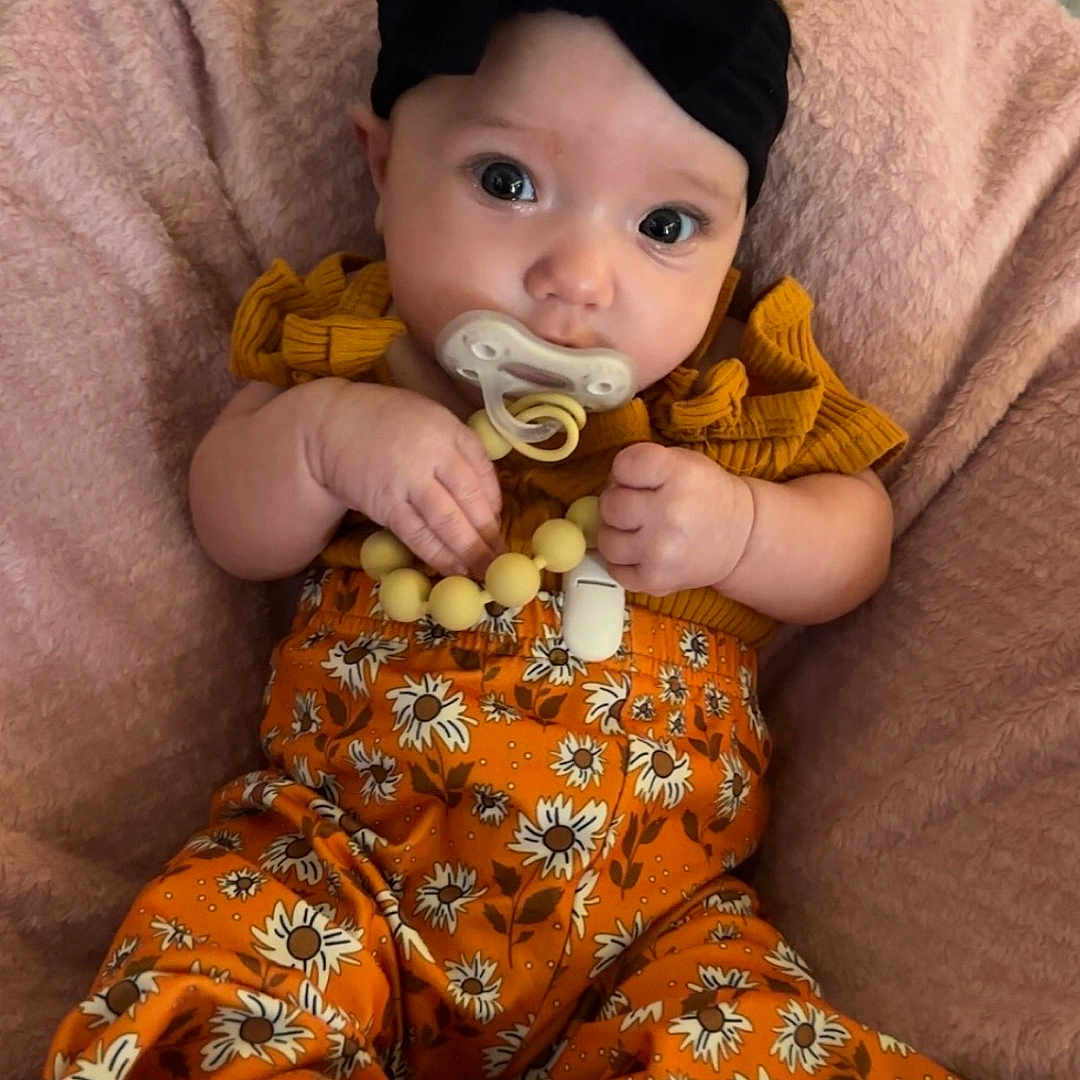 Raivyn is registered to the contest to win money with this photo: baby, black_bow, blanket, child, clothing, cute, floral_pattern, headband, indoor, infant, orange_pants, pacifier, person, pink_blanket, portrait, sitting, small_hands, soft_texture, wide_eyes, yellow_top