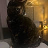 cat, black_cat, pet, animal, feline, sitting, portrait, green_eyes, indoors, cozy, warm_lighting, golden_lights, bokeh, home, furniture, cushion, dark_fur, whiskers, pointed_ears, nighttime