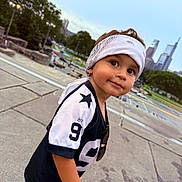 Emiliano is registered to the contest to win money with this photo: child, toddler, bandana, jersey, number_9, sideways_glance, outdoor, concrete, city_skyline, trees, greenery, casual_clothing, short_sleeves, curious_expression, daylight, urban, portrait, smile, young, playful
