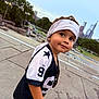 child, toddler, bandana, jersey, number_9, sideways_glance, outdoor, concrete, city_skyline, trees, greenery, casual_clothing, short_sleeves, curious_expression, daylight, urban, portrait, smile, young, playful