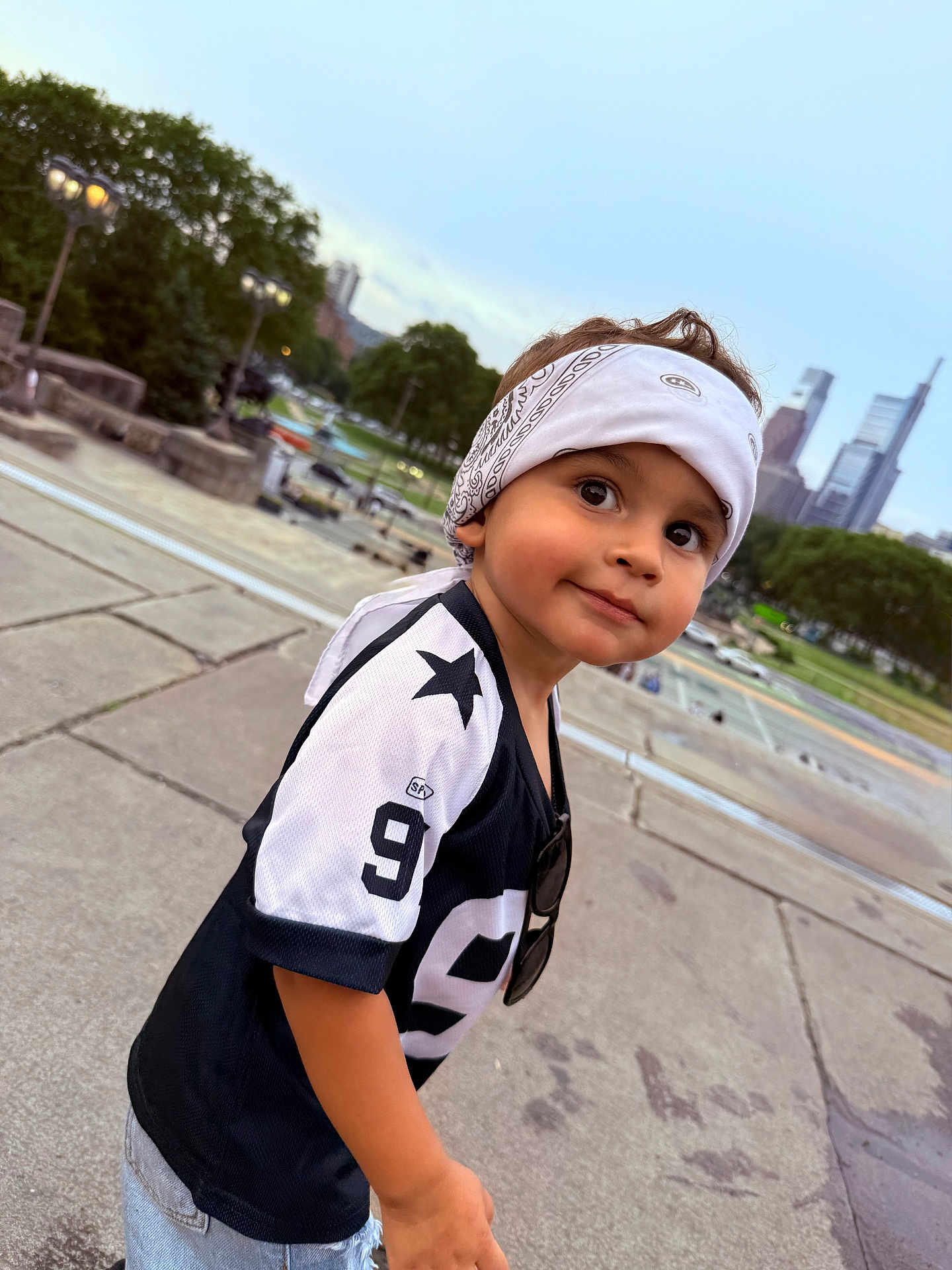 Emiliano is registered to the contest to win money with this photo: child, toddler, bandana, jersey, number_9, sideways_glance, outdoor, concrete, city_skyline, trees, greenery, casual_clothing, short_sleeves, curious_expression, daylight, urban, portrait, smile, young, playful