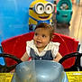 Thea is registered to the contest to win money with this photo: toddler, child, toy_car, red_seat, steering_wheel, minion, background, stroller, indoor, play_area, smiling, clothing, short_hair, blue, yellow, plastic, seat, happy, person, baby