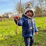 Lucas participe au concours pour gagner de l'argent avec cette photo : child, toddler, blue_jacket, winter_hat, stick, outdoor, grass, nature, exploring, playful, casual_clothing, boots, cold_weather, daylight, smiling, young_child, curious, standing, park, fun