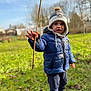 child, toddler, blue_jacket, winter_hat, stick, outdoor, grass, nature, exploring, playful, casual_clothing, boots, cold_weather, daylight, smiling, young_child, curious, standing, park, fun