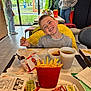 Léo a rejoint le concours — aidez-le/la à gagner de superbes lots ! child, boy, smiling, fast_food, chicken_nuggets, french_fries, drink, table, restaurant, yellow_jacket, casual_clothing, indoor, window, person, food, tray, napkin, sauce, happy, meal