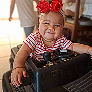 Ohana participe au concours pour gagner de l'argent avec cette photo : baby, child, toy_car, headband, red_bow, striped_shirt, smile, indoor, person, face, hand, bracelet, steering_wheel, blurred_background, adult, furniture, home, play, cute, happy