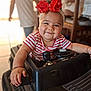 baby, child, toy_car, headband, red_bow, striped_shirt, smile, indoor, person, face, hand, bracelet, steering_wheel, blurred_background, adult, furniture, home, play, cute, happy
