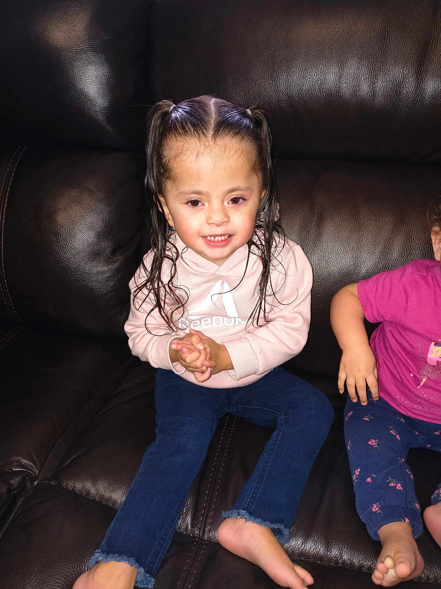 Denise is registered to the contest to win money with this photo: child, girl, pigtails, wet_hair, pink_hoodie, blue_jeans, smile, sitting, leather_couch, indoor, barefoot, casual_clothing, two_children, happy, portrait, young, person, candid, family, cozy