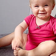 Tilly is registered to the contest to win money with this photo: baby, child, infant, smiling, happy, portrait, indoor, seated, cross_legged, pink_outfit, carpet, remote_control, television_remote, home, floor, blonde_hair, blue_eyes, face, barefoot, adorable