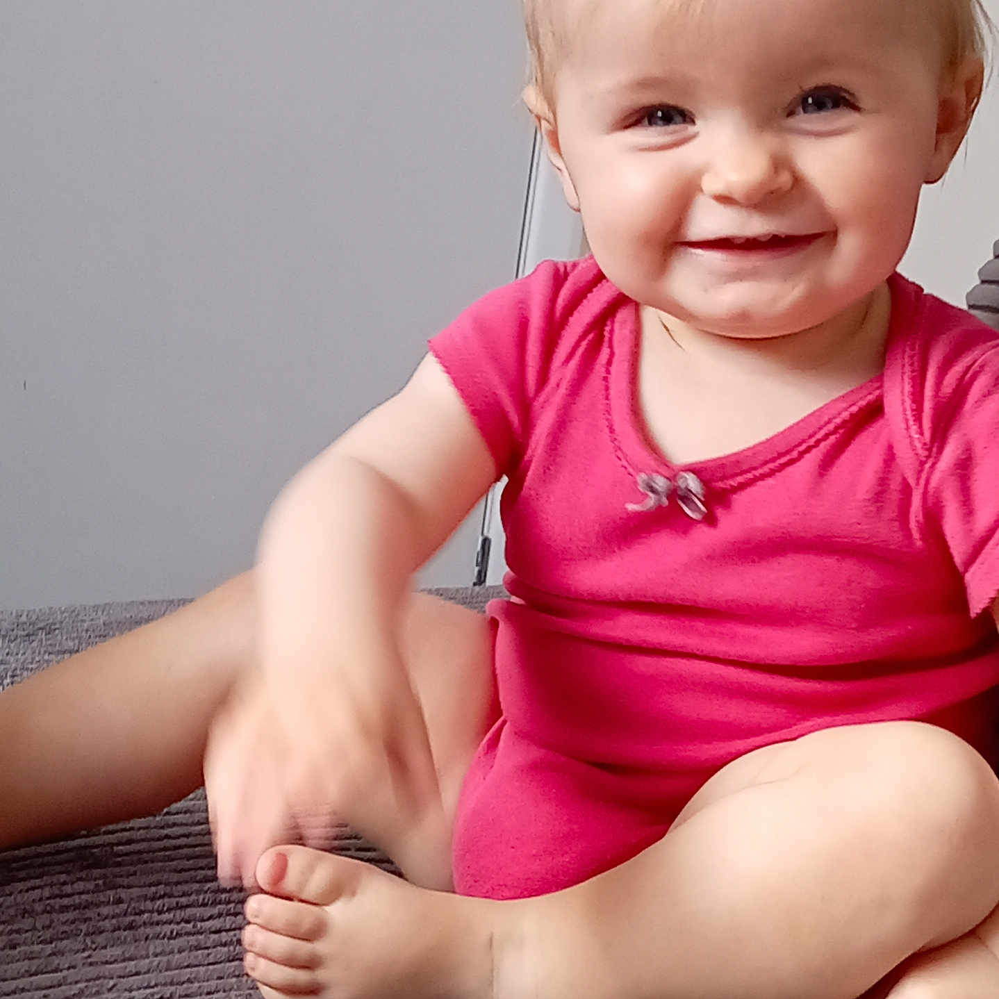 Tilly is registered to the contest to win money with this photo: baby, child, infant, smiling, happy, portrait, indoor, seated, cross_legged, pink_outfit, carpet, remote_control, television_remote, home, floor, blonde_hair, blue_eyes, face, barefoot, adorable