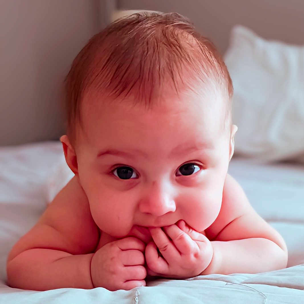 Liliana is registered to the contest to win money with this photo: baby, bed, blanket, chubby_cheeks, close_up, curious, cute, expression, face, hands_in_mouth, head, indoors, infant, lying_down, person, portrait, skin, sleeping_area, soft_lighting, young_child
