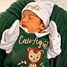 newborn, baby, sleeping, green_outfit, knitted_blanket, lion_embroidery, white_mittens, white_hat, cozy, cute, infant, portrait, soft_lighting, indoors, child, peaceful, warm, snuggled, clothing, baby_fashion