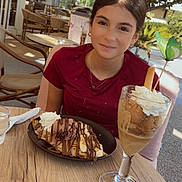 Alyssa a rejoint le concours — aidez-le/la à gagner de superbes lots ! girl, dessert, crepes, chocolate, whipped_cream, iced_coffee, glass, table, outdoor_cafe, chair, tree, leafy, sunlight, smile, face, person, necklace, red_shirt, food, drink
