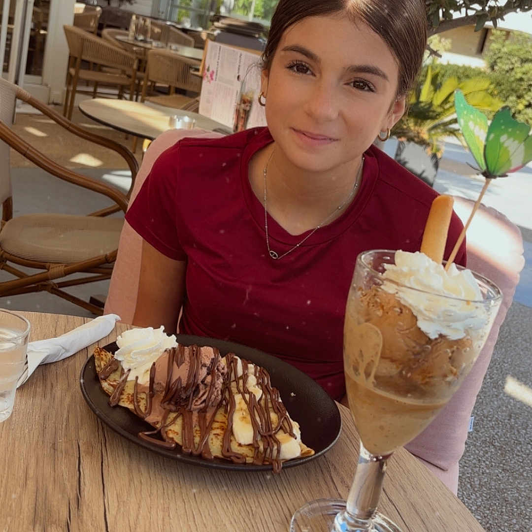 Alyssa a rejoint le concours — aidez-le/la à gagner de superbes lots ! chair, chocolate, crepes, dessert, drink, face, food, girl, glass, iced_coffee, leafy, necklace, outdoor_cafe, person, red_shirt, smile, sunlight, table, tree, whipped_cream