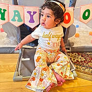 Mahi is registered to the contest to win money with this photo: baby, banner, barefoot, celebration, child, colorful, curly_hair, cute, floor, floral_pattern, headband, indoor, pants, playing_area, playpen, portrait, shirt, sitting, toddler, wooden_floor