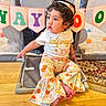 baby, banner, barefoot, celebration, child, colorful, curly_hair, cute, floor, floral_pattern, headband, indoor, pants, playing_area, playpen, portrait, shirt, sitting, toddler, wooden_floor