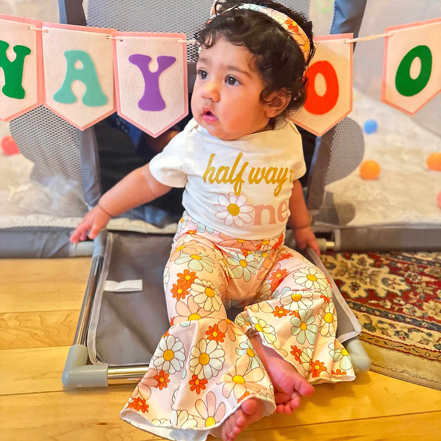 Mahi is registered to the contest to win money with this photo: baby, banner, barefoot, celebration, child, colorful, curly_hair, cute, floor, floral_pattern, headband, indoor, pants, playing_area, playpen, portrait, shirt, sitting, toddler, wooden_floor