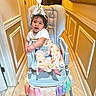 Mahi is registered to the contest to win money with this photo: baby, birthday_hat, celebration, child, colorful, curly_hair, decorations, floor_tile, floral_pants, footwearless, half_birthday, happy, high_chair, indoor, portrait, safety_belt, smiling, tassels, wall_paneling, white_shirt