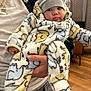 baby, infant, child, onesie, hat, elephant_pattern, hand, person, indoor, wood_floor, jacket, gold_chains, holding, cute, warm_clothing, face, eyes, expression, casual, portrait