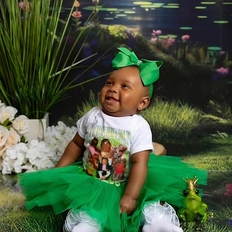 Aurra is registered to the contest to win money with this photo: baby, tutu, green_bow, smiling, toy_frog, crown, flowers, grass, pond, lilypads, white_tights, sitting, happy, portrait, headband, cute, outdoor_scene, child, playful, costume