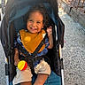 Cairo Rosario joined the competition — help win amazing prizes! toddler, smiling, stroller, child, curly_hair, yellow_bib, red_ball, blue_shirt, shorts, outdoor, sidewalk, sunlight, shadow, metal_fence, happy, baby_toy, seatbelt, portrait, person, urban