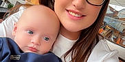 Parker is registered to the contest to win money with this photo: baby, brick_wall, cafe, child, clothing, face, glasses, hair, happy, holding, indoor, light, person, portrait, seated, smile, sweatshirt, tattoo, window, woman