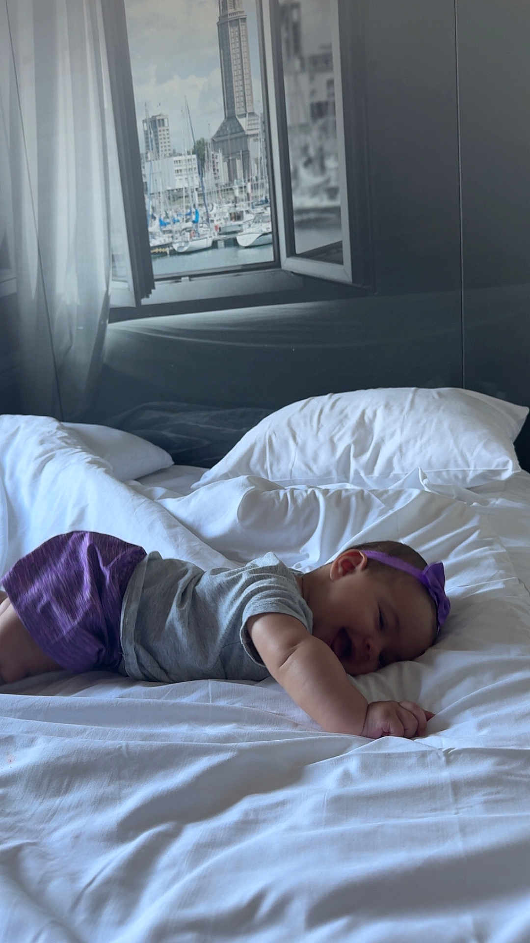 Kamila participe au concours pour gagner de l'argent avec cette photo : baby, bed, white_bed, pillow, purple_headband, purple_shorts, gray_shirt, smiling, happy, infant, indoor, window, curtain, harbor, boats, cityscape, soft_light, sleeping, child, portrait