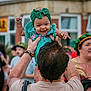 baby, child, smiling, green_headband, green_dress, adult, hands, outdoor, people, building, window, crowd, celebration, happy, portrait, person, festival, daylight, clothing, headwear
