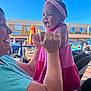 baby, child, woman, outdoor, smiling, holding, pink_dress, headband, laughter, happy, person, sunny, daylight, building, umbrella, car, stroller, seating, people, blue_sky
