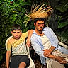 child, young_man, outdoor, tropical, jungle, plants, foliage, portrait, sitting, chairs, basket_hat, sunglasses, casual_clothing, yellow_shirt, white_shirt, shorts, watch, bracelet, serious_expression, photography