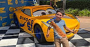 Mikael joined the competition — help win amazing prizes! blue_sky, boy, canopy, checkered_floor, child, dinoco, fun, hedge, outdoor, posing, race_car, shorts, smiling, sneakers, stage_lights, sunny, theme_park, tree, tshirt, yellow_car