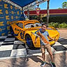 child, boy, smiling, posing, yellow_car, dinoco, race_car, outdoor, sunny, blue_sky, canopy, stage_lights, checkered_floor, shorts, tshirt, sneakers, tree, hedge, theme_park, fun