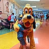 child, mascot, pluto, hug, indoor, people, crowd, cartoon, sketches, wall_art, floor, colorful, smile, shorts, sneakers, backpack, stroller, ceiling, lighting, fun