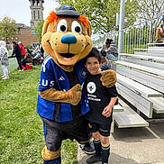 Mikael joined the competition — help win amazing prizes! child, mascot, costume, sports_jersey, outdoor, grass, bleachers, spectators, tree, smile, boy, person, event, daylight, casual_clothing, athletic_shorts, black_shirt, green_socks, concrete_path, friendly