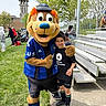 child, mascot, costume, sports_jersey, outdoor, grass, bleachers, spectators, tree, smile, boy, person, event, daylight, casual_clothing, athletic_shorts, black_shirt, green_socks, concrete_path, friendly