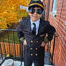 child, young_person, pilot, costume, uniform, aviator_hat, sunglasses, portrait, posing, outdoor, balcony, brick_wall, autumn, fall_leaves, trees, fence, window, formalwear, necktie, smiling